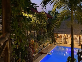 Zona de piscina con una estructura de techo de paja, rodeada de exuberante vegetación y una terraza de madera.