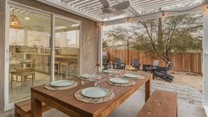Un espacio de comedor al aire libre con una mesa de madera preparada para cuatro personas, rodeada de sillas, y un porche cubierto con ventilador de techo.