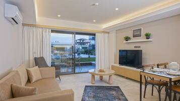 A modern living room with a sofa, a coffee table, and a TV. There is a balcony with a view of buildings and greenery.