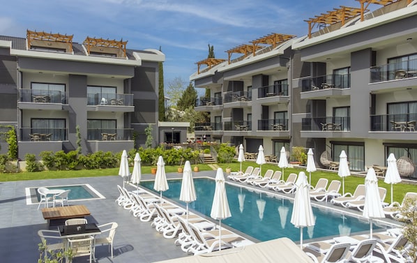 A modern hotel with multiple balconies, a swimming pool with sun loungers, and a well-maintained garden.