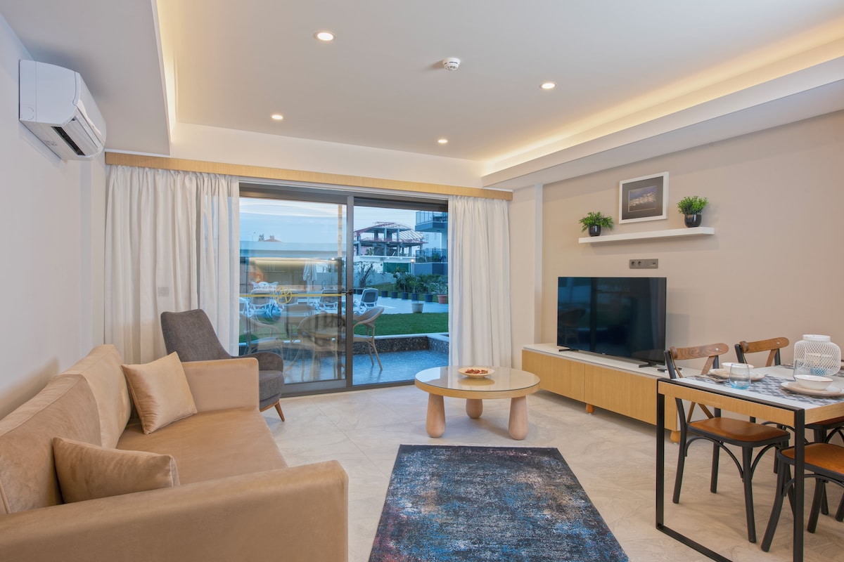 A modern living room with a sofa, a coffee table, and a TV. There is a balcony with a view of buildings and greenery.