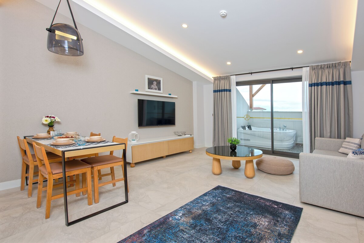 A modern living room with a dining area, a sofa, a TV, and a view of a balcony with a jacuzzi.