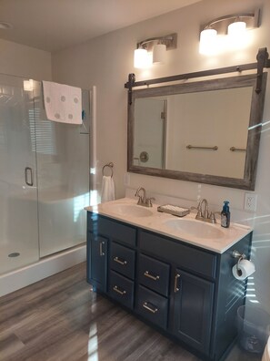 Une salle de bain moderne dotée d’une douche à parois de verre, d’un grand miroir avec des appliques murales et de meubles sombres comprenant un meuble-lavabo.