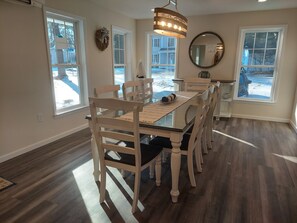 Un espace repas comprenant une table en verre, des chaises en bois, un lustre et de grandes fenêtres donnant sur l’extérieur.