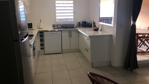 A compact kitchen with white cabinets, a dishwasher, a microwave, and a refrigerator. There is a window with blinds, a sink, and a small fruit bowl on the counter. - Magnificent apartment 2 km from the beach (Le Gosier)