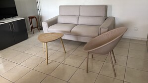 A living room with a sofa, a small round table, a wicker chair, a TV on a cabinet, and a floor lamp. - Magnificent apartment 2 km from the beach (Le Gosier)