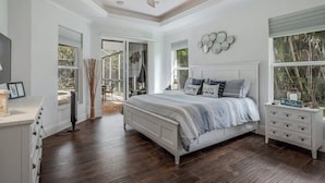 A bedroom with a large bed, a ceiling fan, a dresser, a nightstand, and a window with a view of trees.