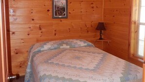 Quarto de cabana de madeira com colcha acolchoada, um quadro na parede e uma mesa de cabeceira com abajur.