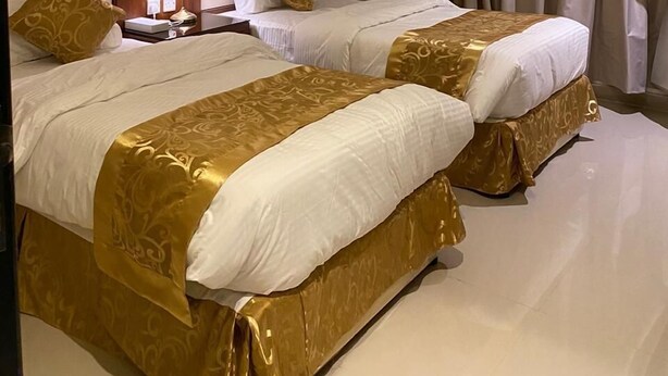A hotel room with two beds, each with a gold and white bedspread, a nightstand with a lamp, and a large window with curtains. - Rihanna Inn APT - Riyadh (Riyadh)