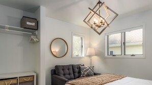 A bedroom with a bed, a grey sofa, a round mirror, a window with blinds, a hanging light fixture, and storage units. - Fish Creek Beach House, Westview: 2 Bed, 2 Bath (Fish Creek)