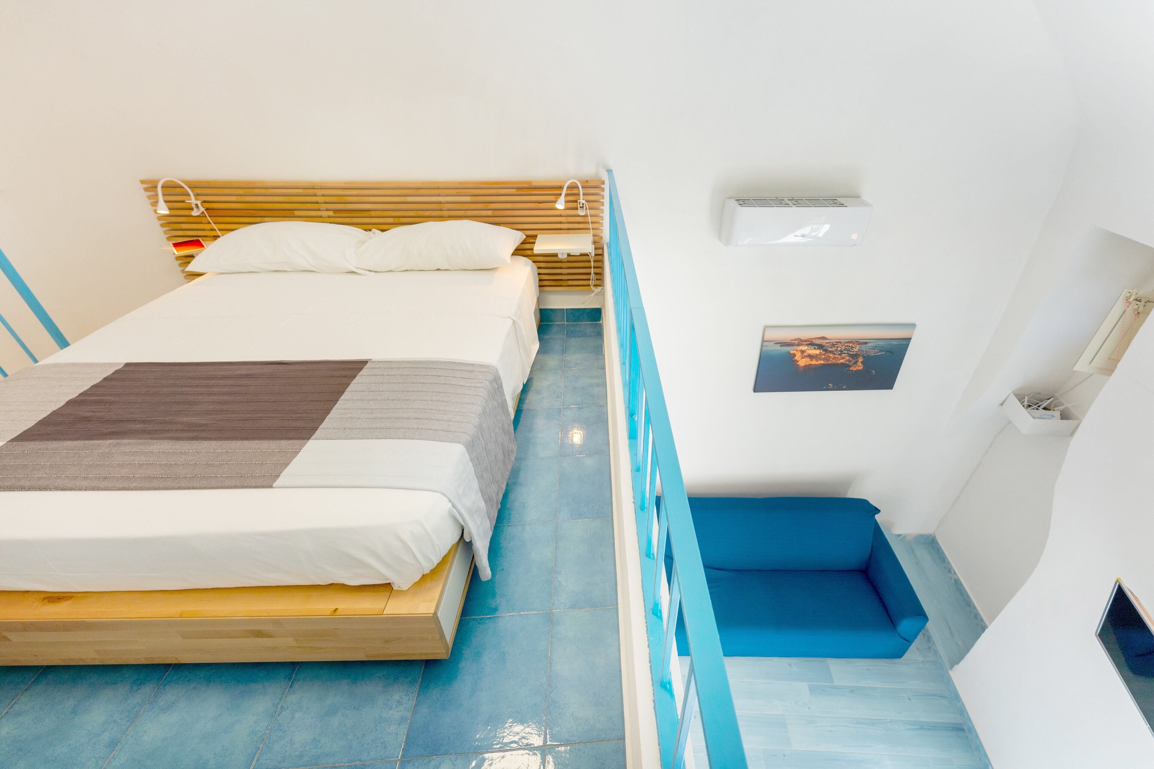 Quarto com cama, um banco azul, uma mesinha e ar condicionado fixado na parede.