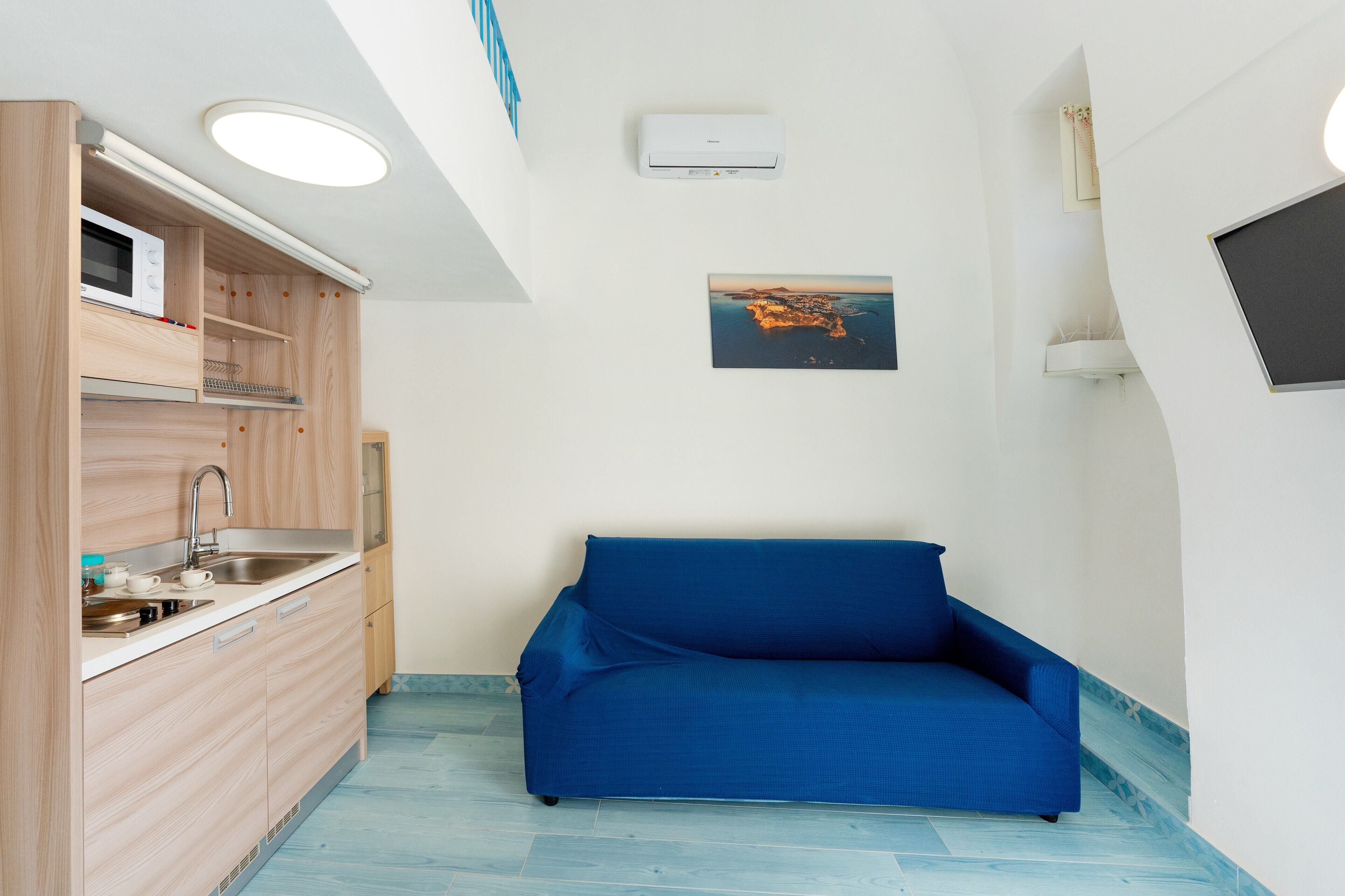 A compact room with a kitchenette, a blue sofa, a ceiling-mounted air conditioner, and a framed picture on the wall.