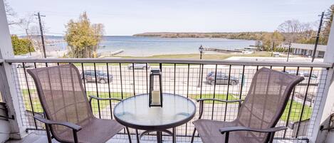 A balcony with two wicker chairs and a glass table, overlooking a waterfront with a parking lot and buildings in the background.