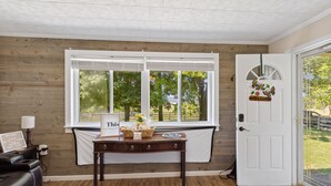 A room with a wooden wall, a white door, a window with blinds, a table with a sign, a lamp, and a bench.