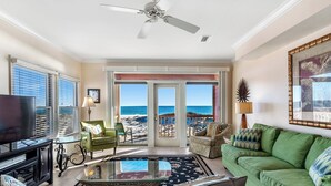 A living room with a green sofa, a glass coffee table, and a television. There is a balcony with a view of the beach.