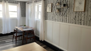 A dining area with a wooden table and chairs, a window with white curtains, and a wall with a tree pattern wallpaper and framed artwork. - Brand new Apt B: 1 Bedroom 1 Bath in the Heart of Downtown Ellsworth (Ellsworth)