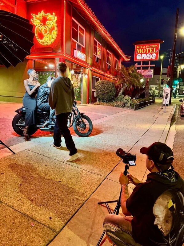 A brightly lit motel with neon signage, a motorcycle parked outside, and a person filming. - Royal Pagoda Motel (Los Angeles)