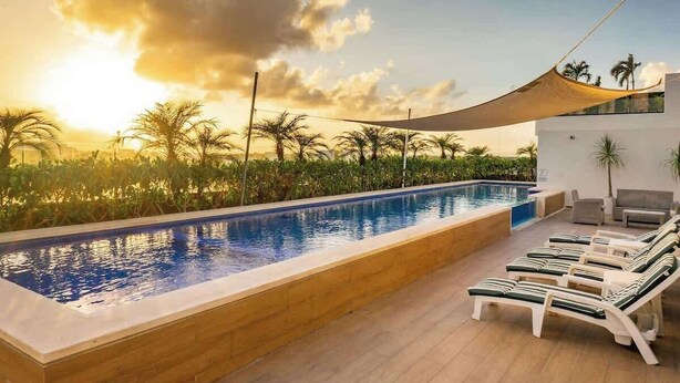 Piscina com espreguiçadeiras, cercada por palmeiras e um pôr do sol.