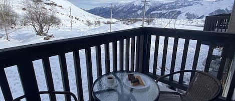 A balcony with a glass table and two wicker chairs, overlooking a snowy mountain landscape.