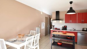 Una cocina compacta con armarios rojos, una lámpara colgante negra y una zona de comedor con sillas blancas y una mesa de madera.
