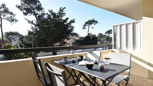 Un balcón con una mesa dispuesta para dos personas, sillas y vistas a árboles y casas.
