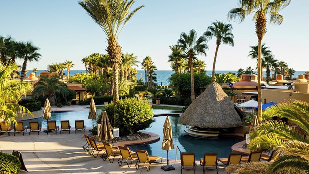 A resort pool area with lounge chairs and umbrellas, surrounded by palm trees and a view of the sea.