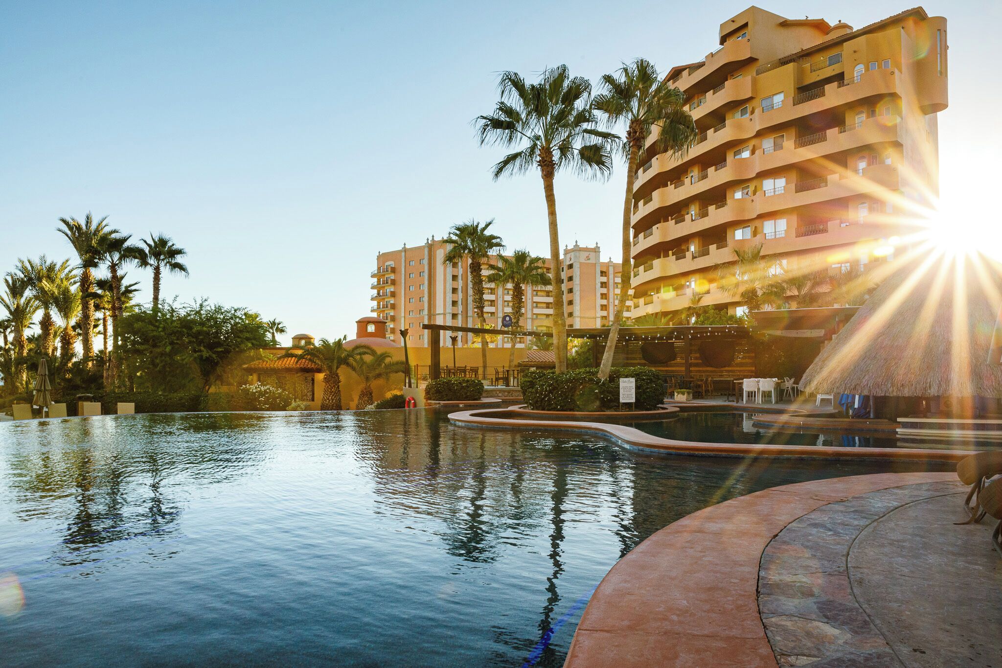 A resort pool area with palm trees, a thatched-roof structure, and a multi-story building in the background.