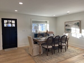 Un coin repas avec une table en bois, six chaises, un tapis, une porte noire, une fenêtre avec des stores et un tableau accroché au mur.