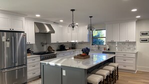 Una cocina moderna con gabinetes blancos, electrodomésticos de acero inoxidable, una isla grande con taburetes de bar e iluminación colgante.