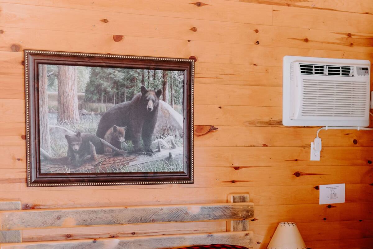 A wooden cabin room with a bed, a framed picture of bears, an air conditioning unit, and a lamp.