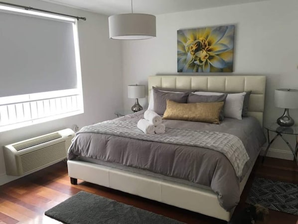 A bedroom with a large bed, a window with blinds, a radiator, and a painting on the wall.