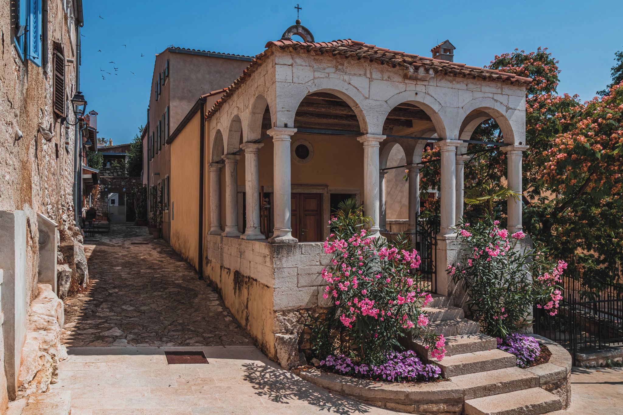 Une rue étroite bordée d’immeubles en pierre, un escalier menant à un bâtiment orné d’arcades et des plantes en fleurs.