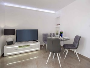 A modern living room with a flat-screen TV, a white entertainment unit, a dining table with chairs, and a small side table with a vase and wine bottles.