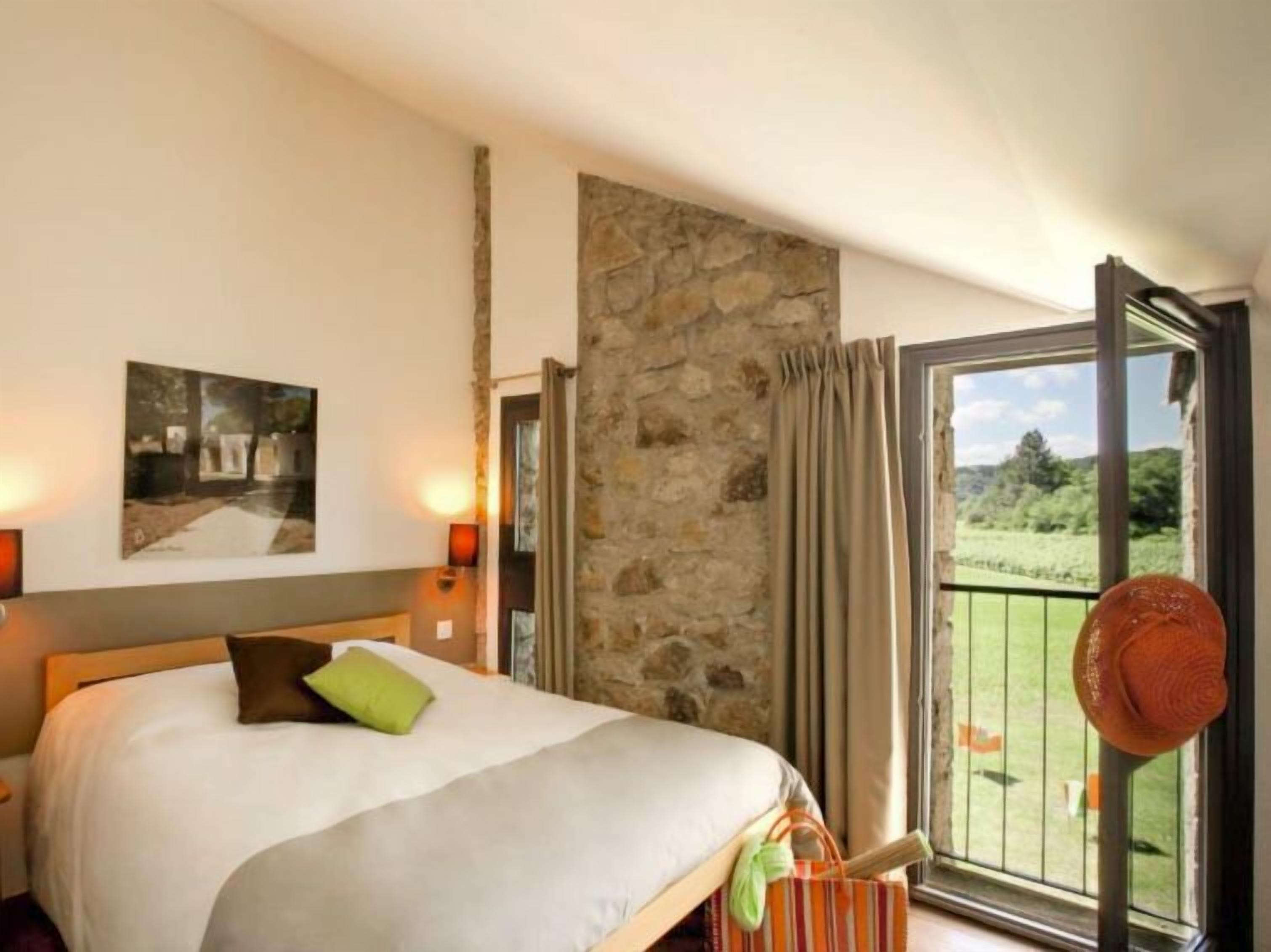 Une chambre d’hôtel avec un lit, un mur en pierre, un balcon donnant sur un paysage verdoyant et un tableau accroché au mur.