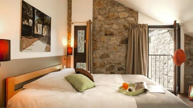 Une chambre avec un mur en pierre, un lit avec des oreillers, une table de chevet avec une lampe et une fenêtre avec des rideaux.