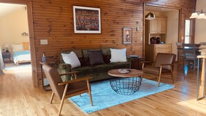A cozy cabin interior with a wooden floor, a green sofa with white cushions, two brown armchairs, a round coffee table, and a kitchen area in the background. - ✶Appalachian Mt Cabin, Mt Views, 3BR 3BA, Fiber Wifi, Family, Hiking, Waterfalls (Unicoi)