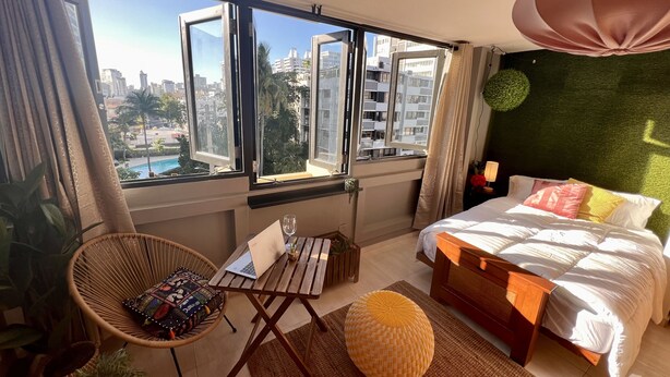 A modern hotel room with a large window, a bed, a chair, and a small table. - Lovely beach studio - High speed internet! (San Juan)