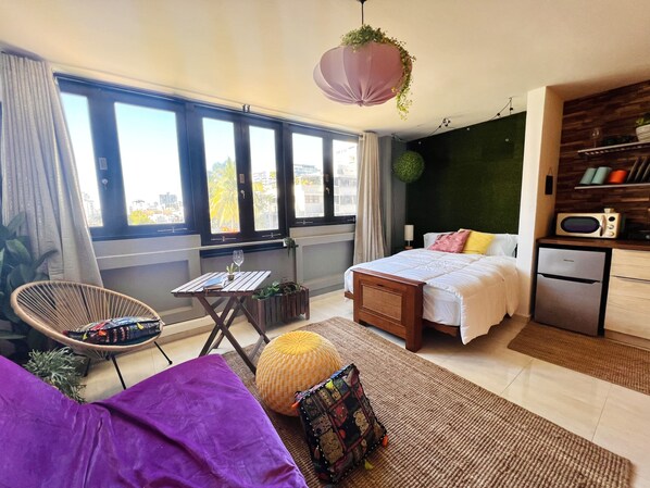 A modern hotel room with a bed, a small kitchenette, a dining area with a table and chair, and large windows offering a city view.
