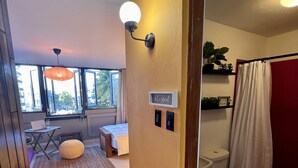 A hotel room with a bed, bedside table, and a small table with a chair. There is a window with a view of the outside, a bathroom with a toilet and a shower curtain, and a hallway with a door leading to another room. - Lovely beach studio - High speed internet! (San Juan)
