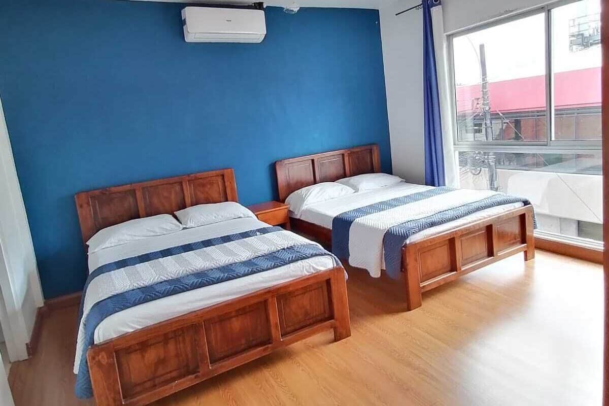 A hotel room with two wooden beds, a blue wall, a window with a view, and an air conditioning unit.