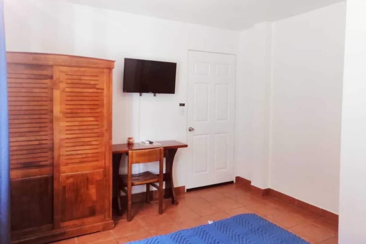 A room with a wooden wardrobe, a desk with a chair, a flat-screen TV mounted on the wall, and a closed door.
