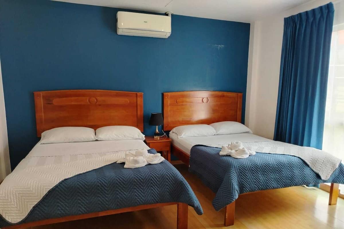 A hotel room with two wooden beds, a blue wall, an air conditioning unit, and a window with blue curtains.