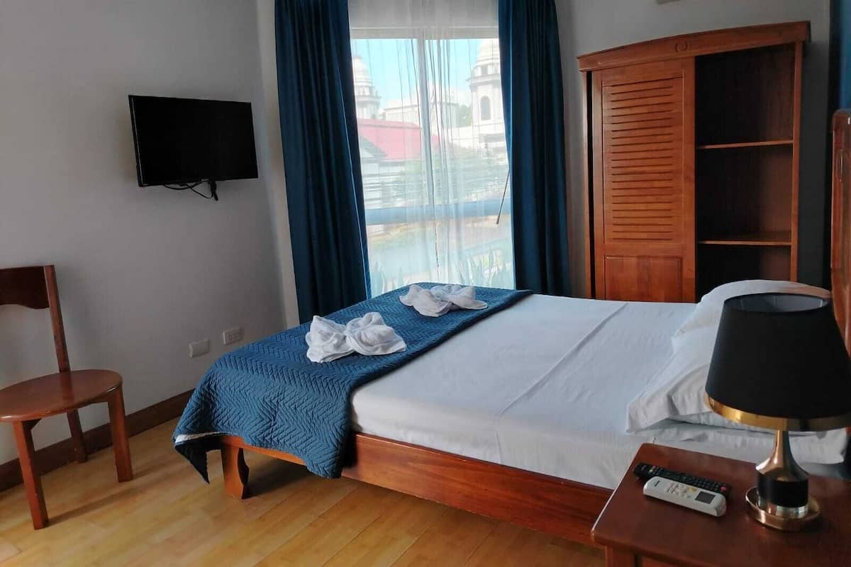 A hotel room with a bed, a TV mounted on the wall, a wooden wardrobe, and a window with curtains.