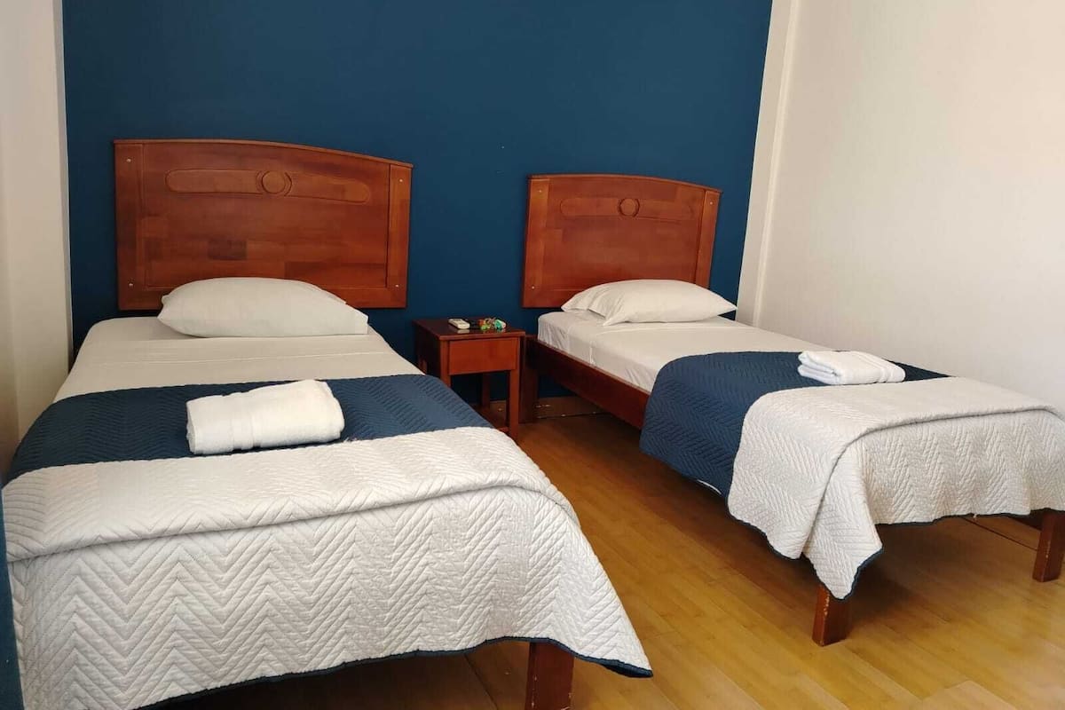 A hotel room with two single beds, each with a wooden headboard, white bedding, and a blue blanket. There is a small bedside table with a lamp and a nightstand with a lamp.