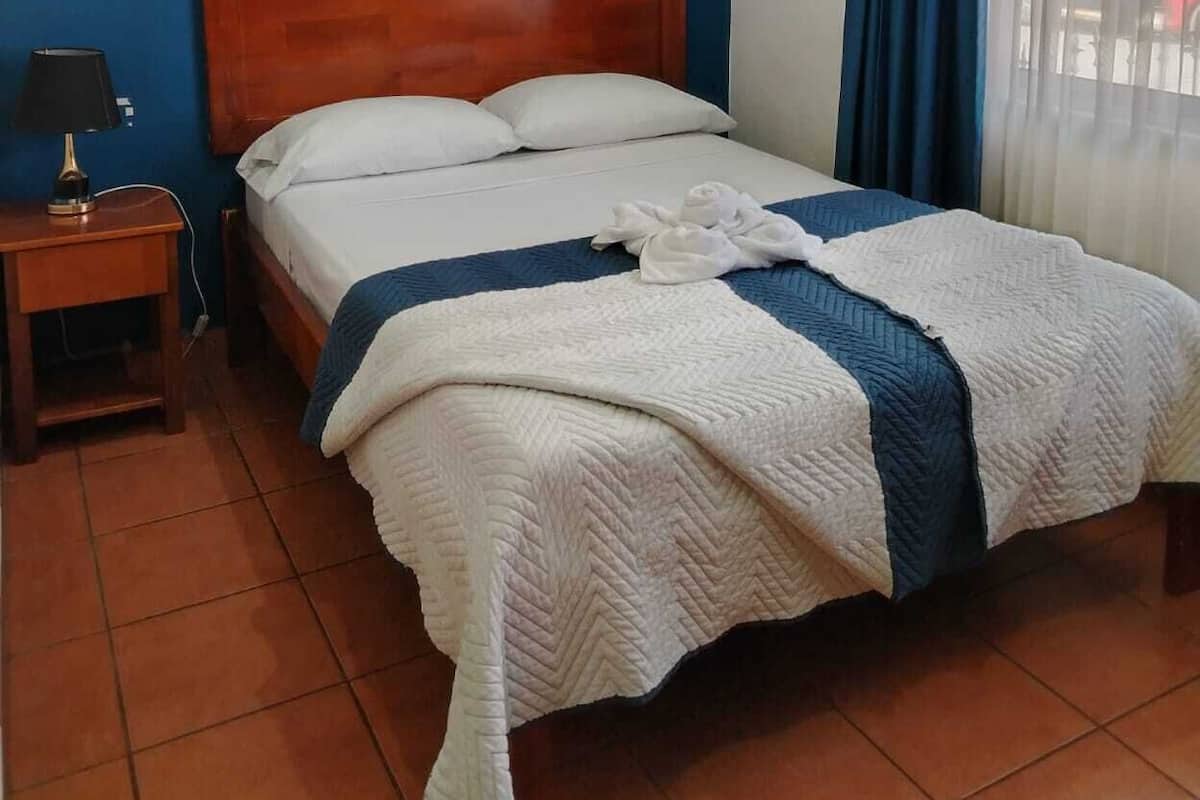 A hotel room with a wooden headboard, a bed with white and blue bedding, an air conditioning unit on the wall, a nightstand with a lamp, and a window with blue curtains.