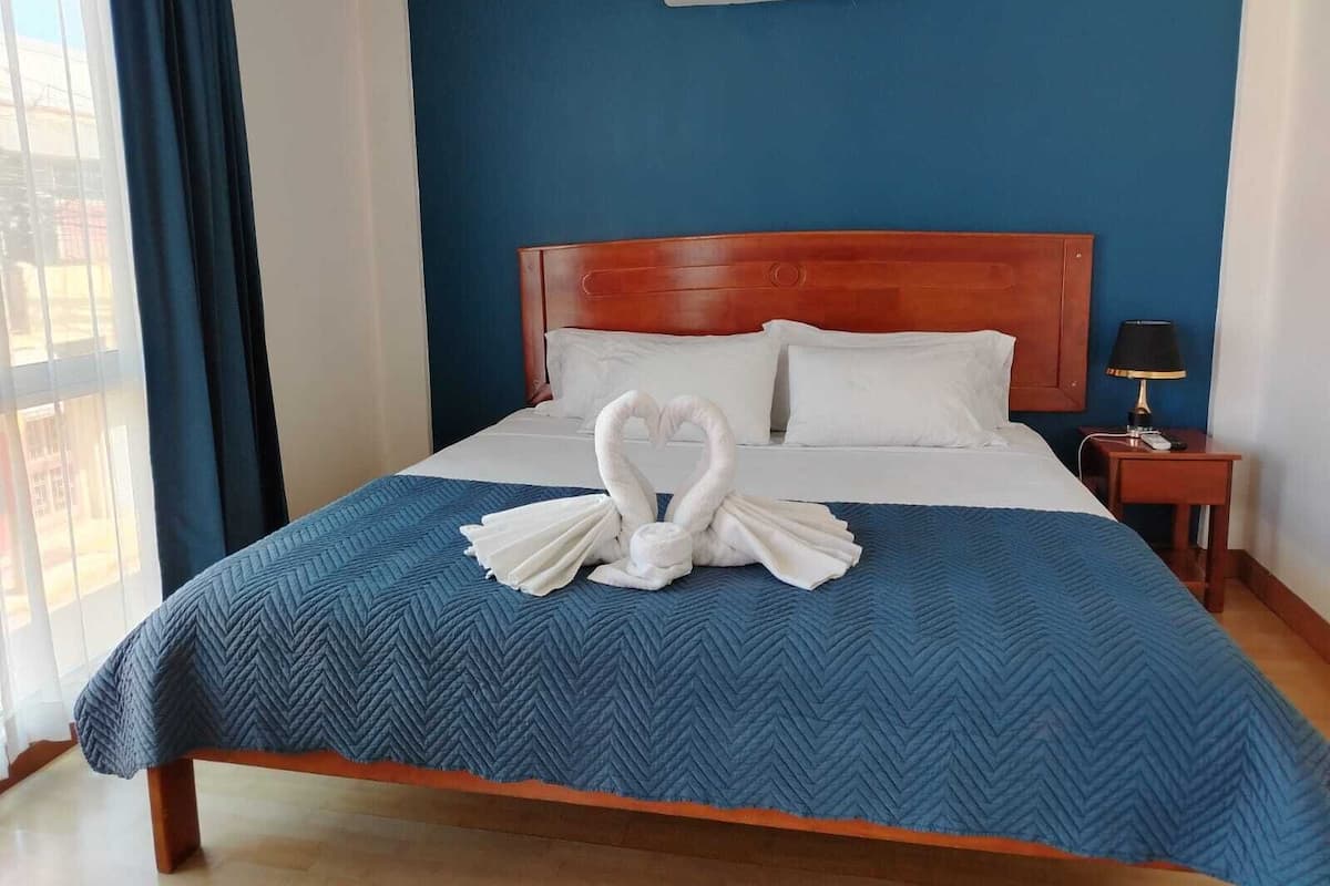 A hotel room with a bed, a wooden headboard, a blue wall, and a window with curtains.