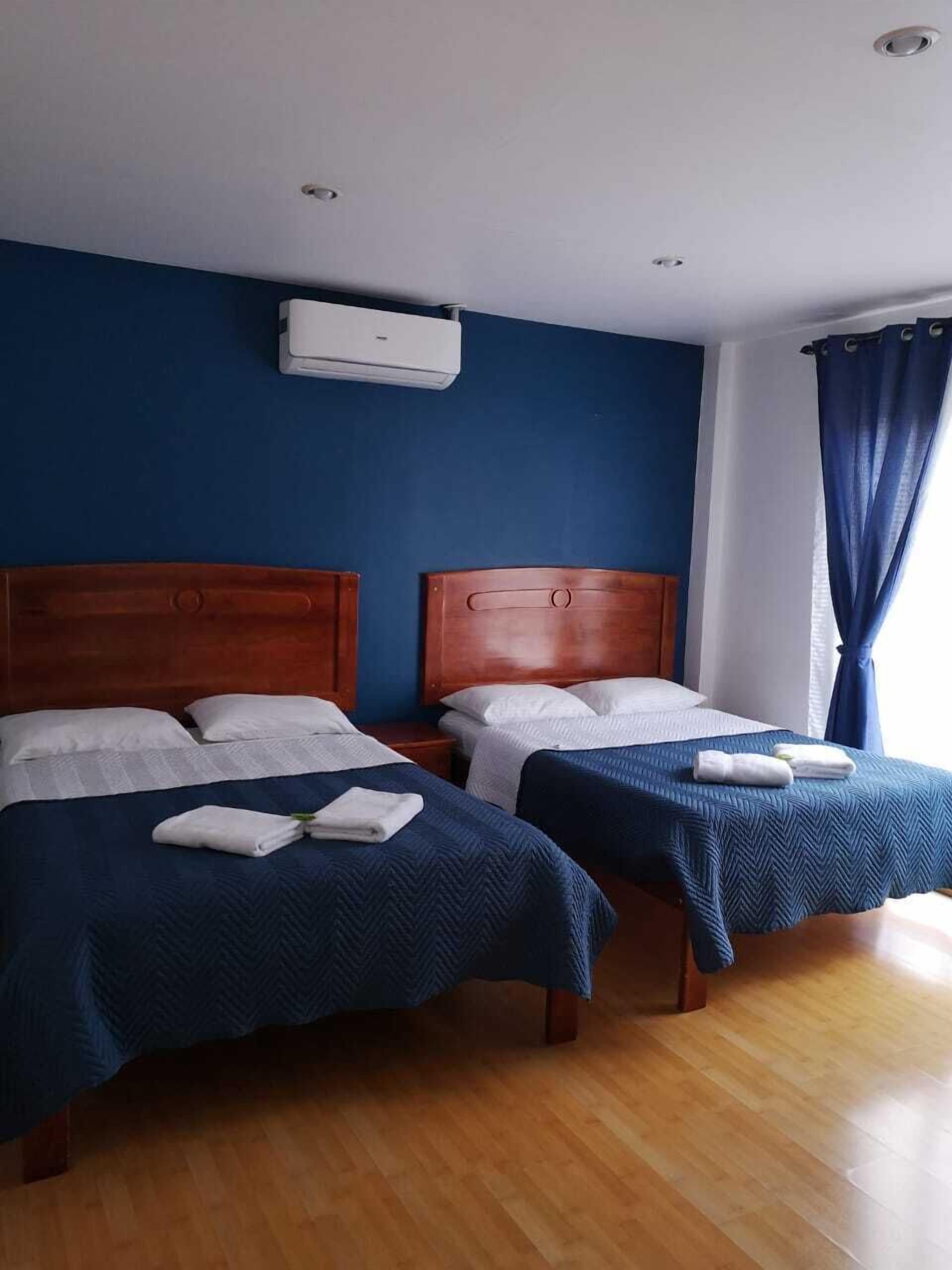 Habitación de hotel con dos camas, cabeceros de madera, ropa de cama blanca, una pared azul y un equipo de aire acondicionado.