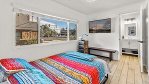 A bedroom with a large bed, a bench, a dresser, and a bathroom with a sink and mirror. - Beaver Street Stone Lodge w/AC, Fully Fenced Yard, W/D, 3 Smart TVs, Speedy WiFi (Flagstaff)