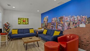 A living room with a blue sofa, red armchair, and a coffee table. There is a bookshelf with books and a painting of apples on the wall. - Beaver Street Stone Lodge w/AC, Fully Fenced Yard, W/D, 3 Smart TVs, Speedy WiFi (Flagstaff)
