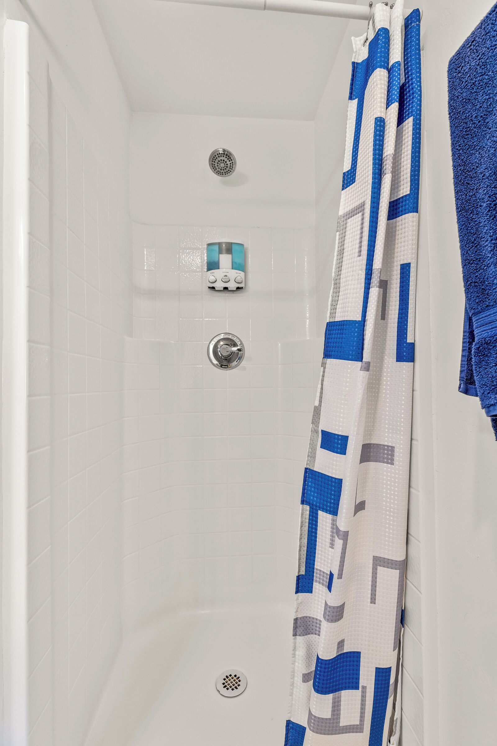 A bathroom with white tiled walls, a shower with a blue and white patterned shower curtain, and a blue towel hanging on the wall.